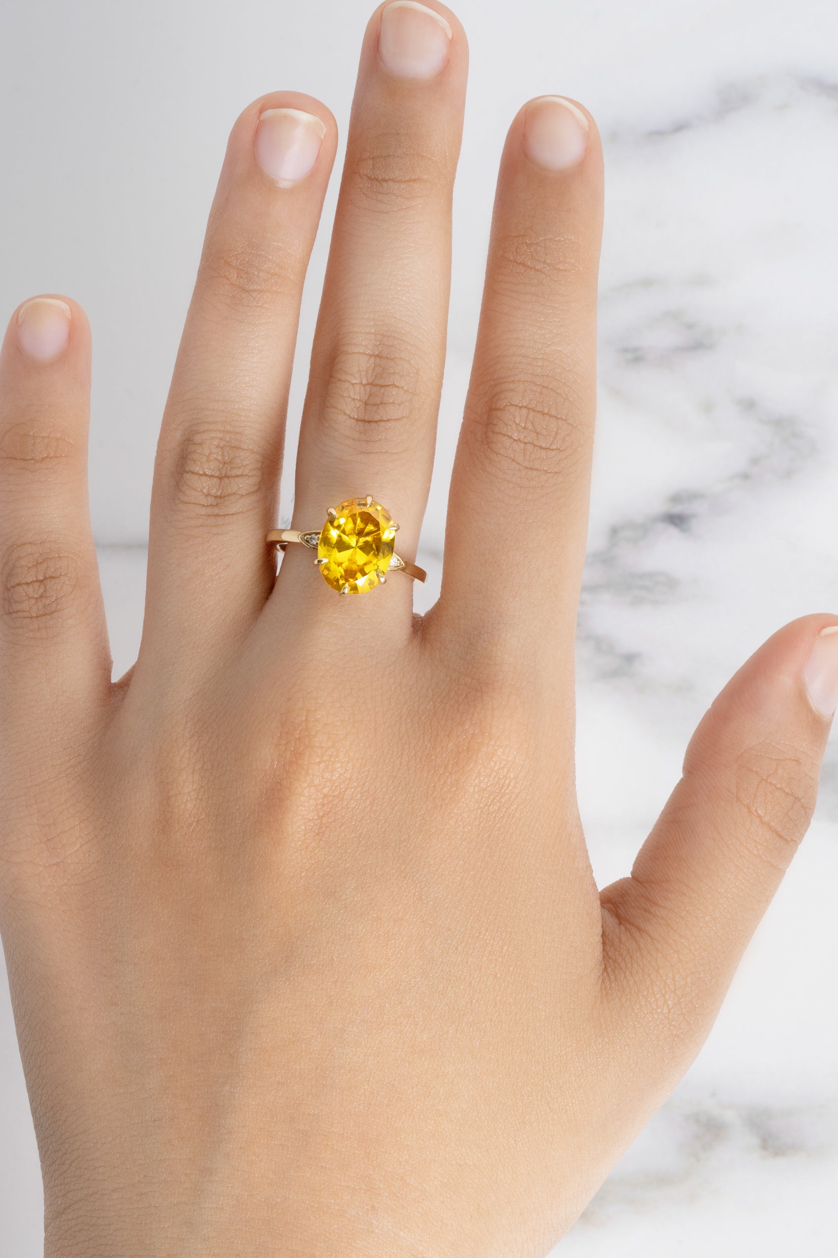 A hand models the Flora Oval Yellow Sapphire Cocktail Ring, showing its brilliant faceted stone and gold band on a marble surface.