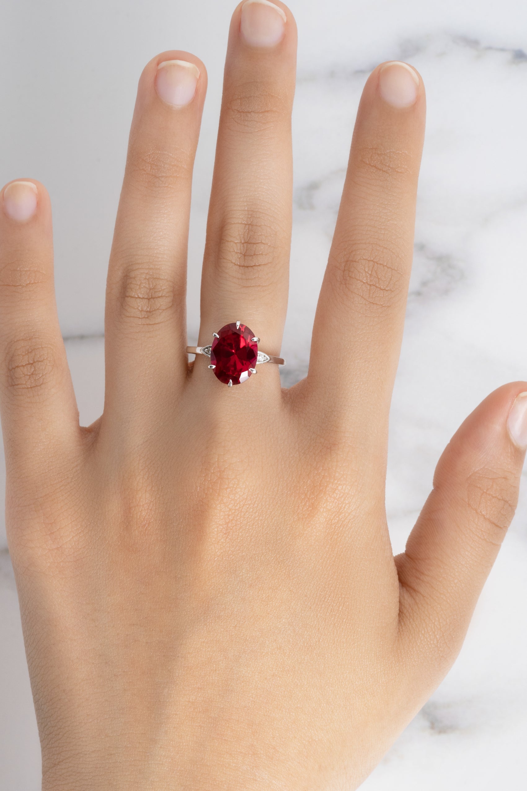 A woman's hand modeling the Flora Oval Ruby Cocktail Ring with diamond accents in 18k white gold against a marble surface.