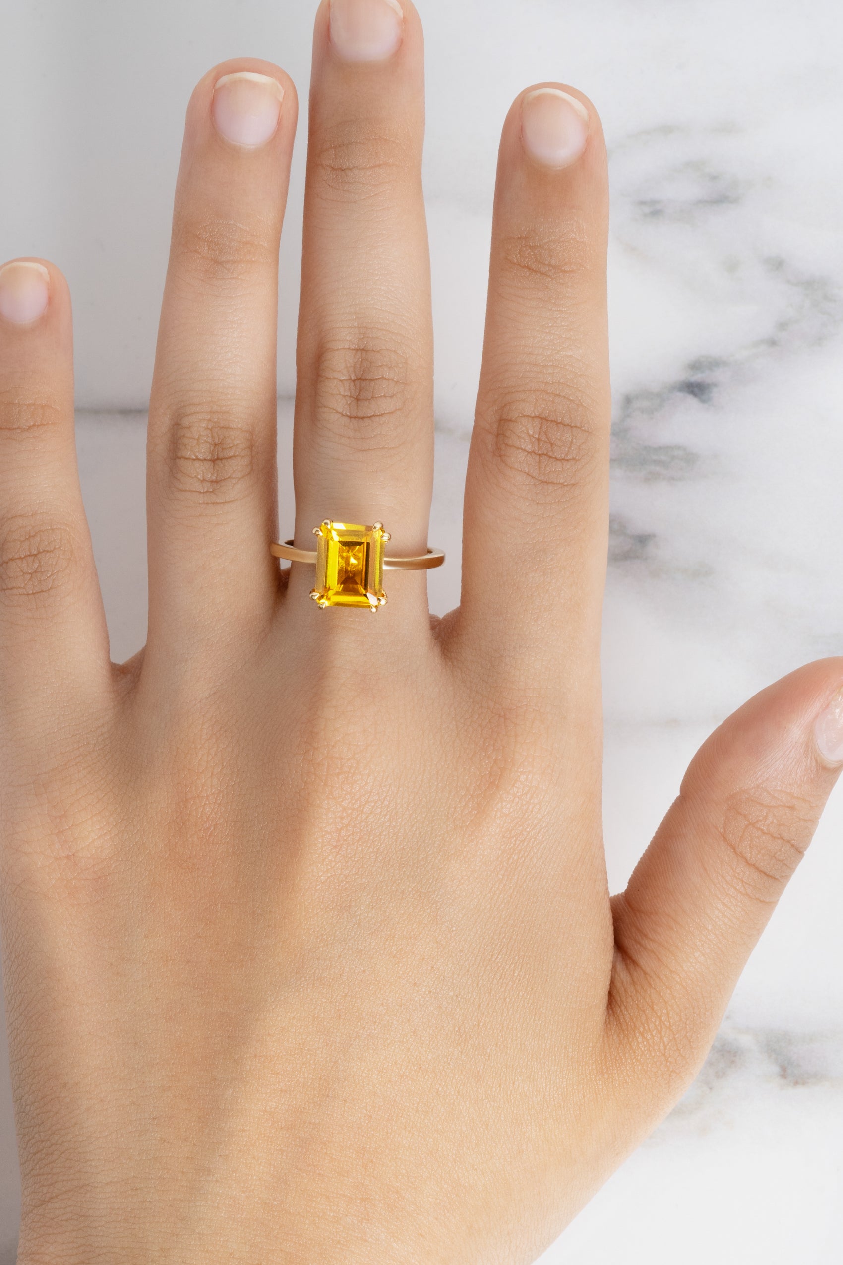 A model's hand wearing the Flora Yellow Sapphire Octad Cocktail Ring with a gold band against a white marble background.