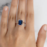 A model's hand wearing the Flora Cocktail Ring with a large, rectangular blue sapphire on a silver-toned band.