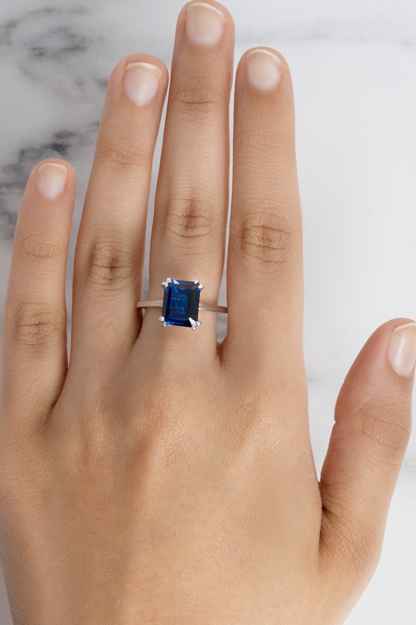 A model's hand wearing the Flora Cocktail Ring with a large, rectangular blue sapphire on a silver-toned band.