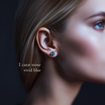 Close-up of a model wearing a 1 carat vivid blue, brilliant round cut diamond stud earring against a dark background.